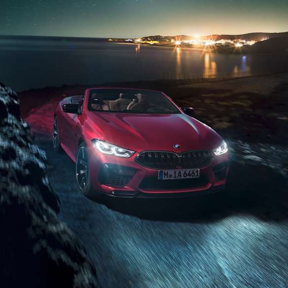BMW M8 Competition Кабриолет, вид спереди с поворотом в три четверти. BMW M8 Competition Кабриолет в Motegi Red металлик с открытым мягким верхом, вид спереди с поворотом в три четверти, езда ночью по прибрежной дороге.