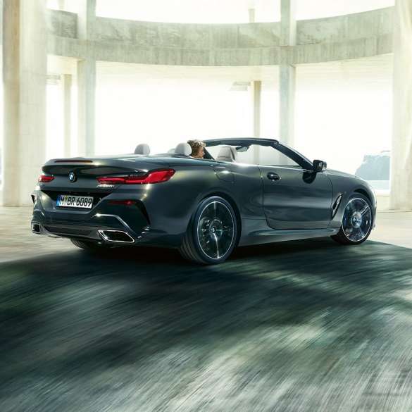 BMW M850i xDrive Кабриолет, вид сзади с поворотом в три четверти. BMW M850i xDrive Кабриолет в Dravit Grey металлик с открытым мягким верхом, вид сзади с поворотом в три четверти, перед бетонным зданием.