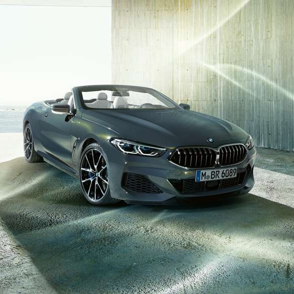 BMW M850i xDrive Кабриолет, вид спереди с поворотом в три четверти. BMW M850i xDrive Кабриолет в Dravit Grey металлик с открытым мягким верхом, вид спереди с поворотом в три четверти, на берегу.