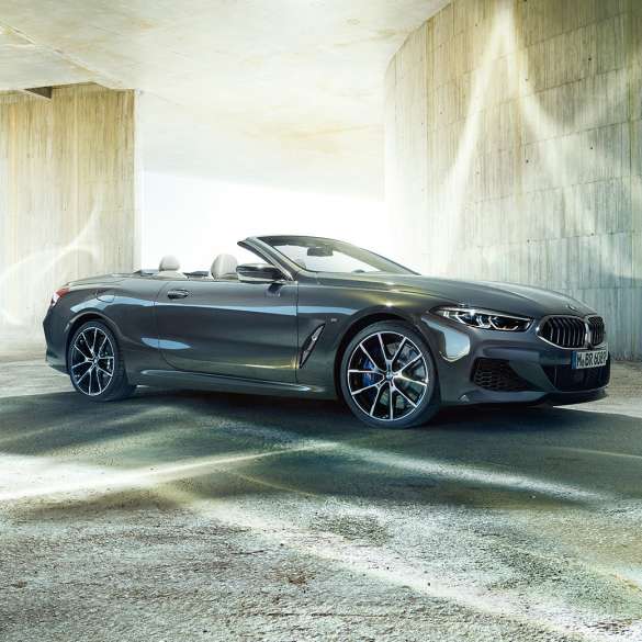 BMW M850i xDrive Кабриолет, вид сбоку с поворотом в три четверти. BMW M850i xDrive Кабриолет в Dravit Grey металлик с открытым мягким верхом, вид сбоку с поворотом в три четверти, перед бетонным зданием.