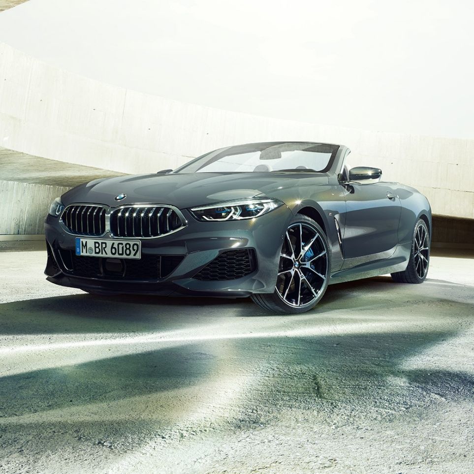 BMW M850i xDrive Кабриолет, вид спереди с поворотом в три четверти. BMW M850i xDrive Кабриолет в Dravit Grey металлик с открытым мягким верхом, вид спереди с поворотом в три четверти, перед бетонным зданием.