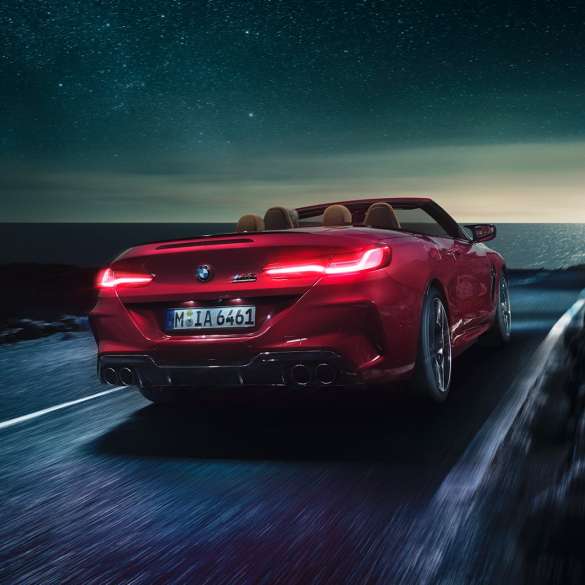 BMW M8 Competition Convertible, three-quarter rear view. BMW M8 Competition Convertible in Motegi Red metallic with open softtop, three-quarter rear view, driving at night on coast road.