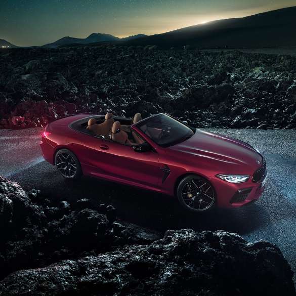 BMW M8 Competition Convertible, three-quarter side view. BMW M8 Competition Convertible in Motegi Red metallic with open softtop, three-quarter side view, standing on asphalt at night.