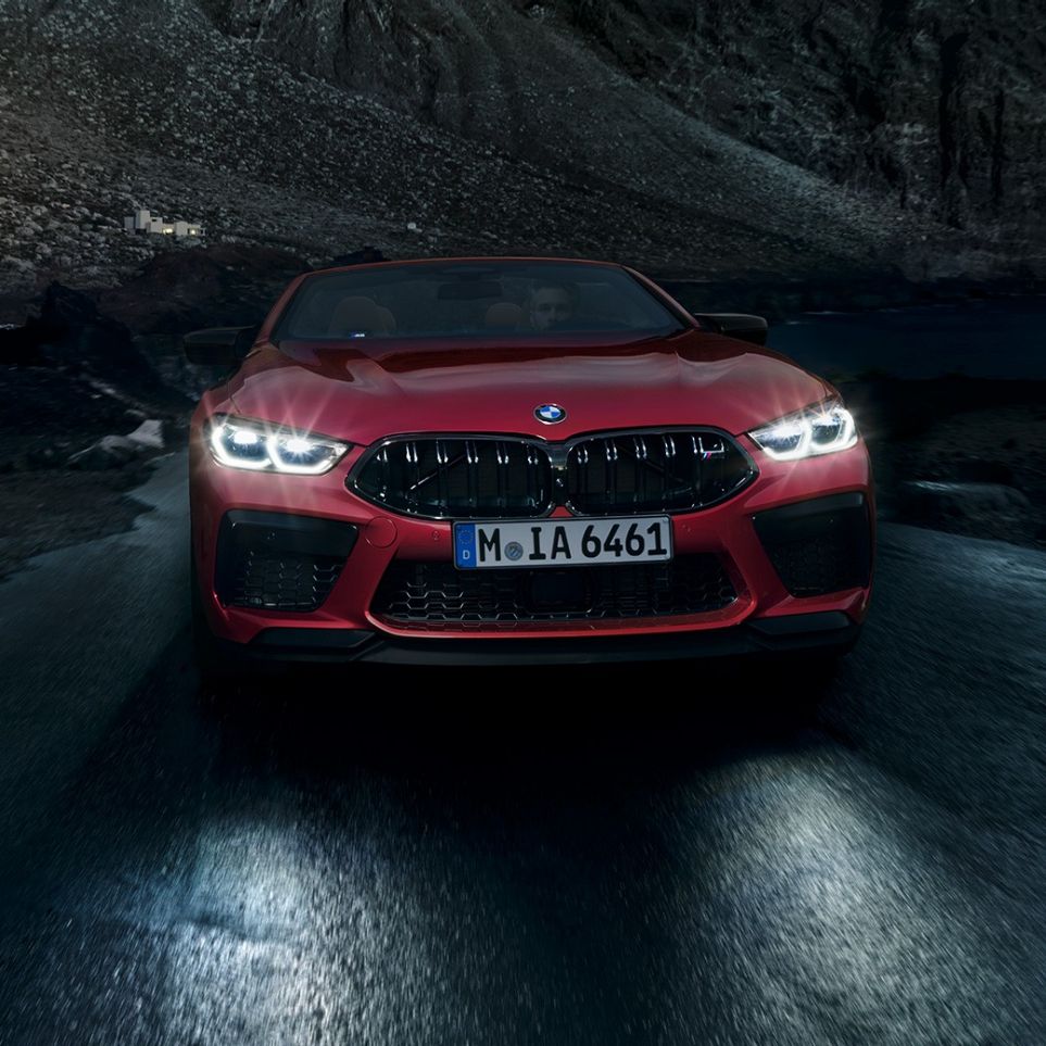 BMW M8 Competition Convertible, front view. BMW M8 Competition Convertible in Motegi Red metallic with shining headlights, front view, driving at night.