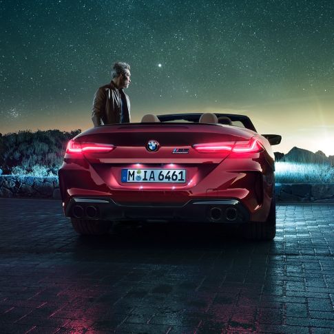 BMW M8 Competition Convertible, rear view. BMW M8 Competition Convertible in Motegi Red metallic with open softtop, rear view, standing with man under starry sky.