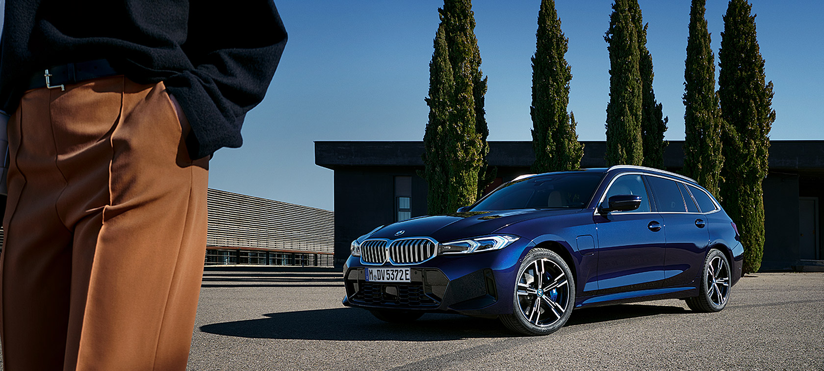 BMW 3 серии Туринг LCI G21 в цвете Tansanit Blue II, вид спереди с поворотом в три четверти на фоне деревьев