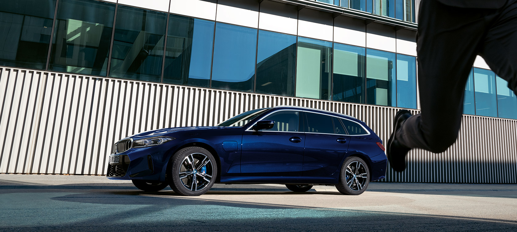 BMW 3 серии Туринг LCI G21, вид сбоку с поворотом в три четверти на фоне здания BMW 3 серии Туринг LCI G21 в цвете Tansanit Blue II, вид сбоку с поворотом в три четверти на фоне здания