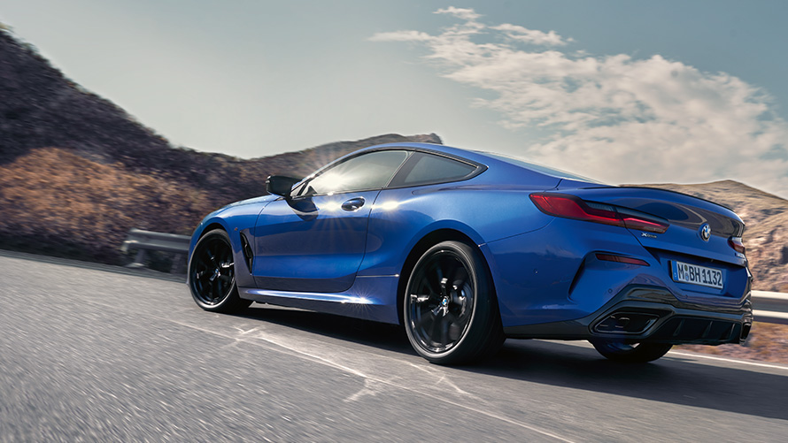BMW 8 серии Coupé G15 LCI (рестайлинг 2022 года) вид сзади с поворотом в три четверти, в движении BMW 8 серии Coupé G15 LCI (рестайлинг 2022 года) в цвете Синий Портимау металлик, вид сзади с поворотом в три четверти, в движении на фоне холмов