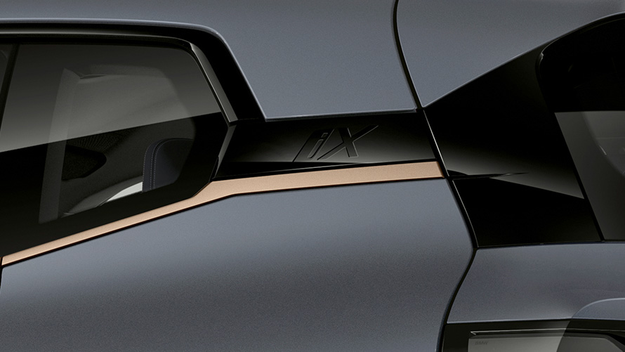 BMW iX M60 i20 2022 электрический SUV, электрический автомобиль в цвете Oxid Grey metallic, крупный план на значок iX рядом с изгибом Хофмайстера с отделкой BMW Individual Exterior Line Titanium Bronze