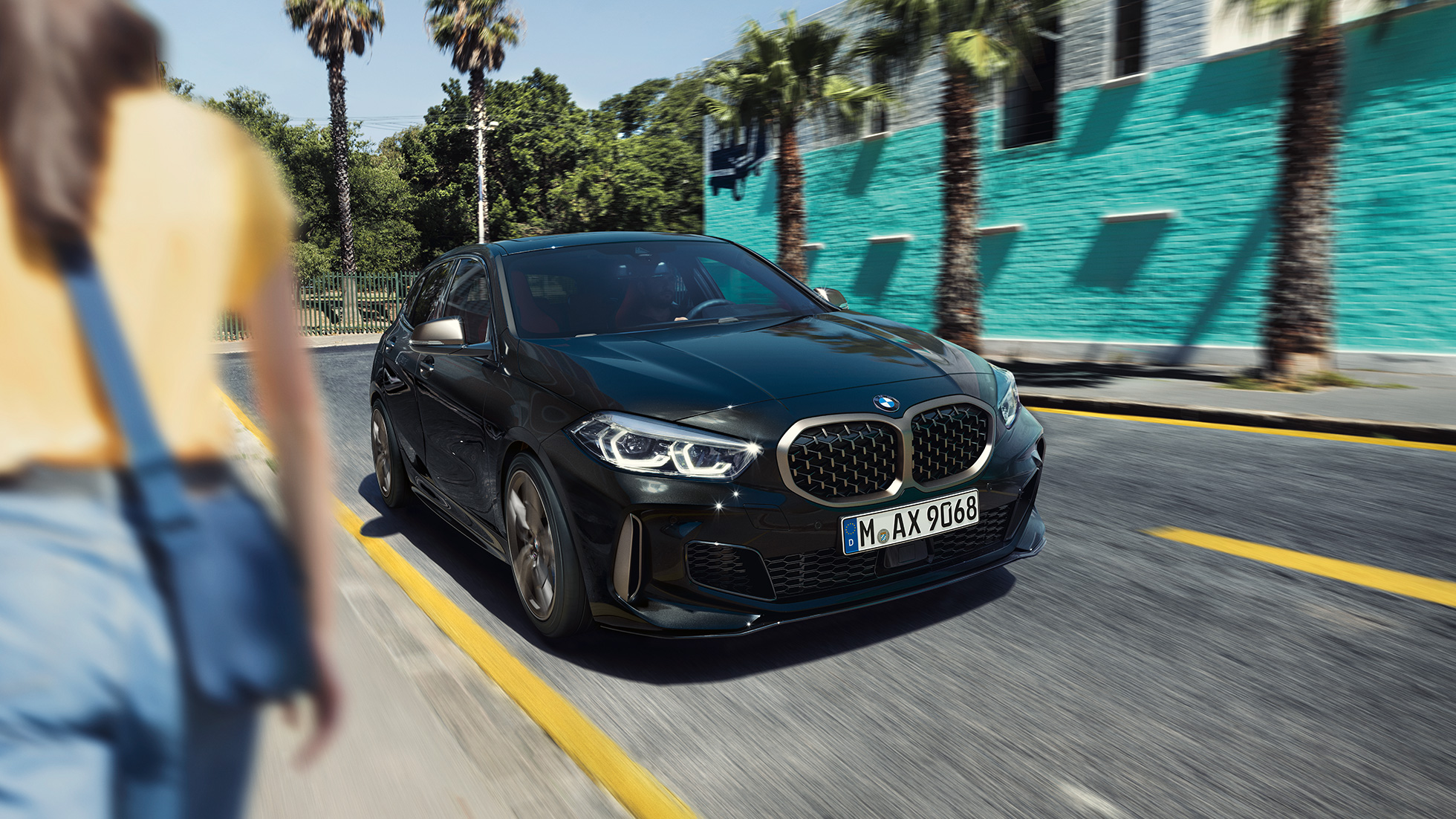 BMW M135i xDrive, вид спереди с поворотом в три четверти. BMW M135i xDrive в Black Sapphire металлик, вид спереди за рулем с поворотом в три четверти с женщиной на фоне.