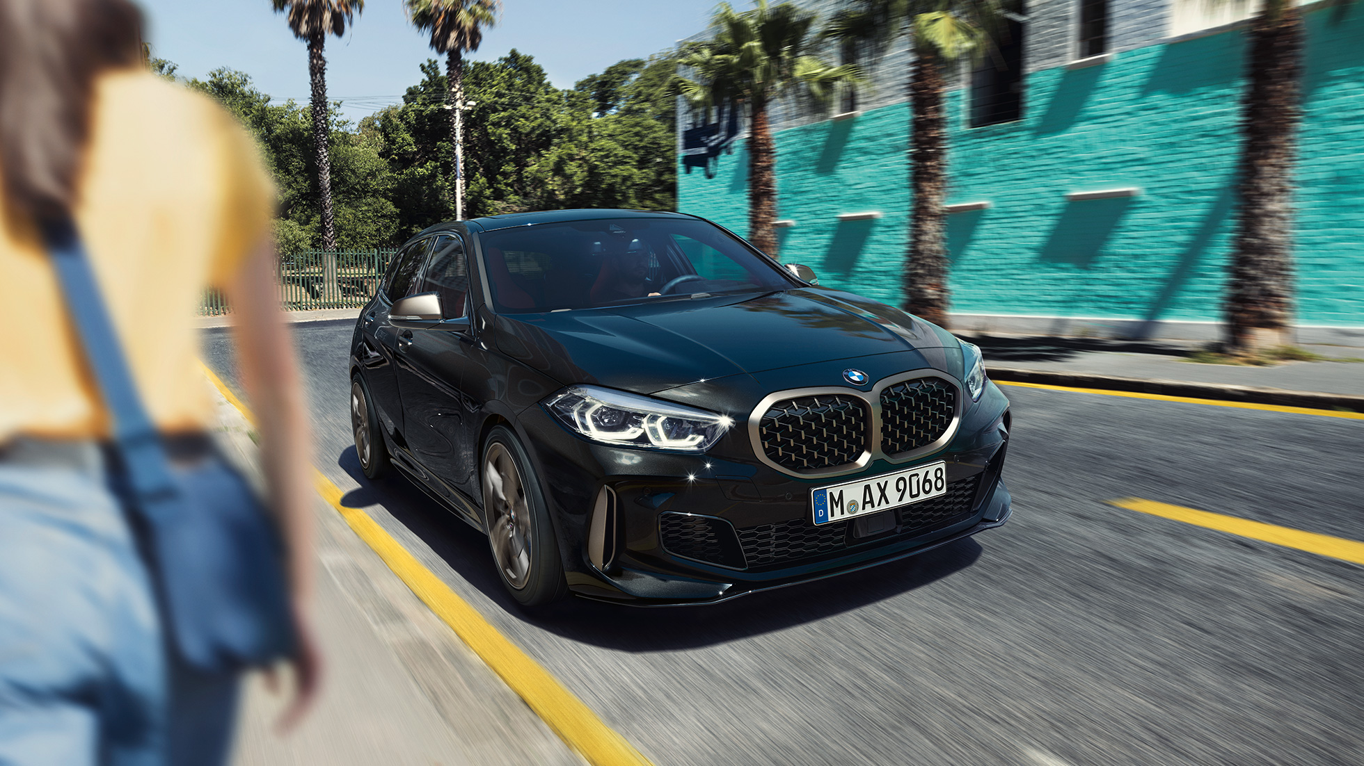 BMW M135i xDrive, вид спереди с поворотом в три четверти. BMW M135i xDrive в Black Sapphire металлик, вид спереди за рулем с поворотом в три четверти с женщиной на фоне.