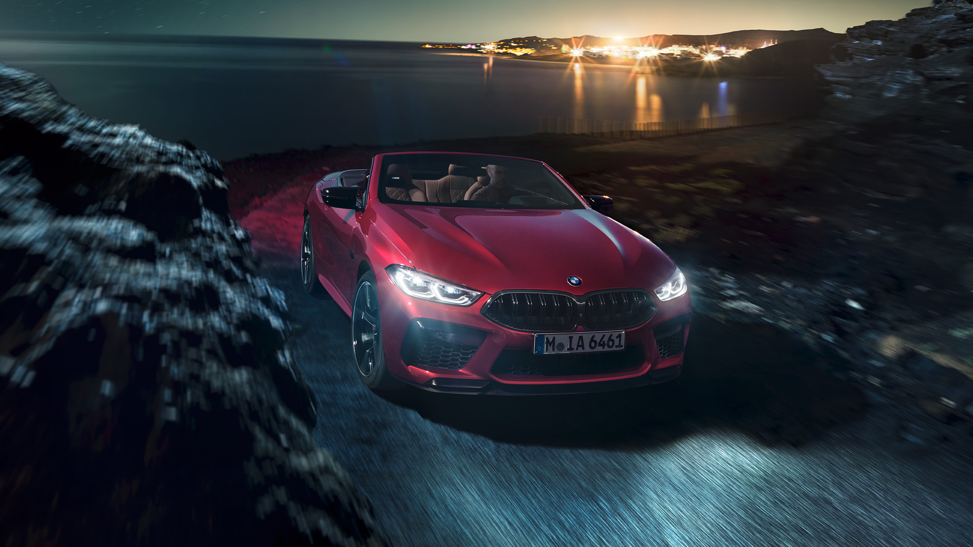 BMW M8 Competition Кабриолет, вид спереди с поворотом в три четверти. BMW M8 Competition Кабриолет в Motegi Red металлик с открытым мягким верхом, вид спереди с поворотом в три четверти, езда ночью по прибрежной дороге.