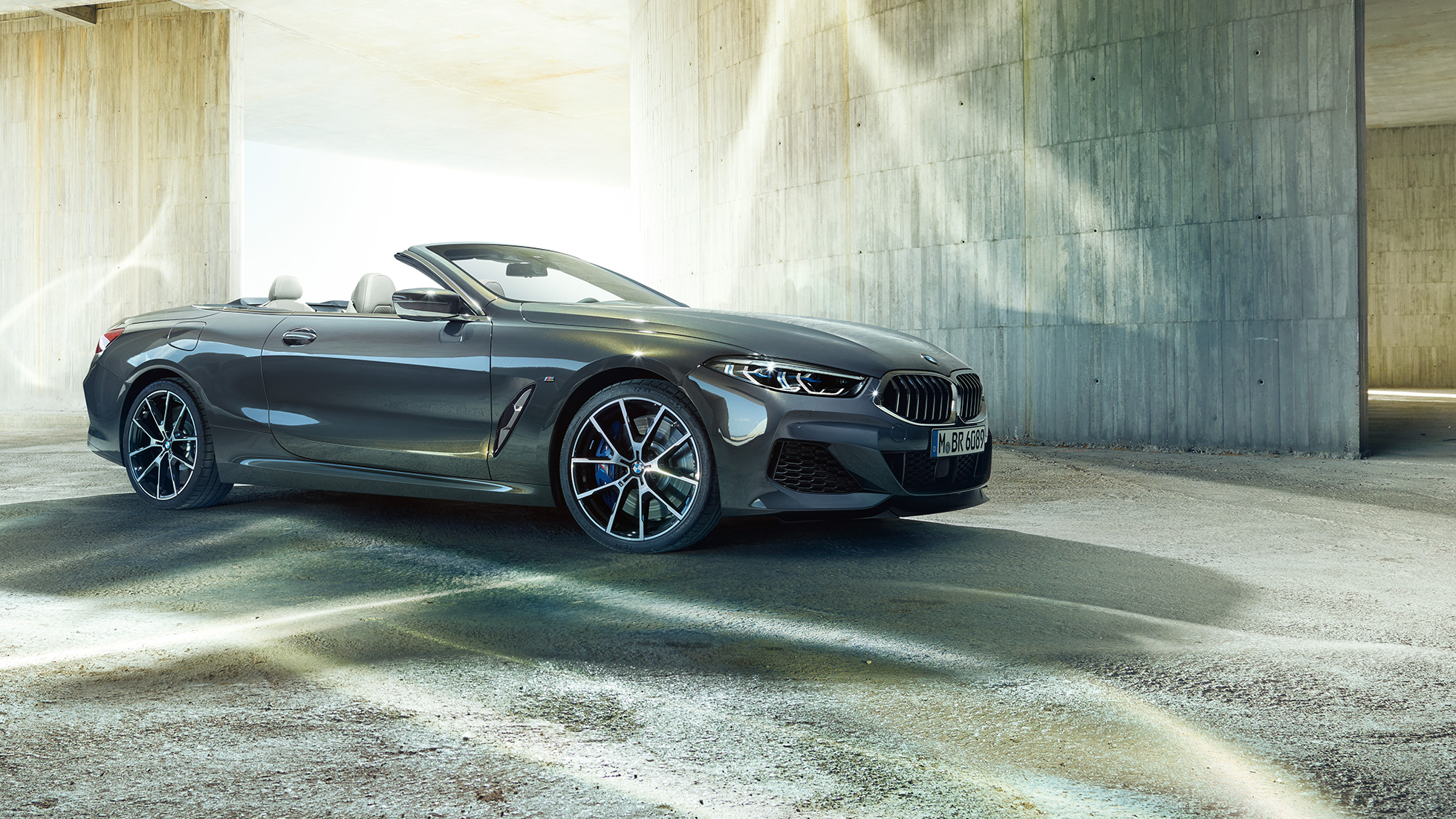 BMW M850i xDrive Кабриолет, вид сбоку с поворотом в три четверти. BMW M850i xDrive Кабриолет в Dravit Grey металлик с открытым мягким верхом, вид сбоку с поворотом в три четверти, перед бетонным зданием.