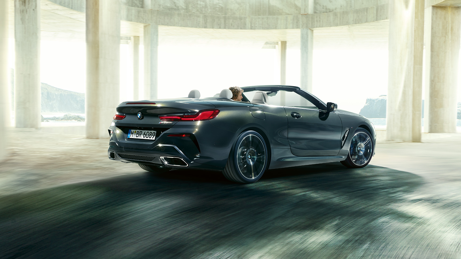 BMW M850i xDrive Кабриолет, вид сзади с поворотом в три четверти. BMW M850i xDrive Кабриолет в Dravit Grey металлик с открытым мягким верхом, вид сзади с поворотом в три четверти, перед бетонным зданием.