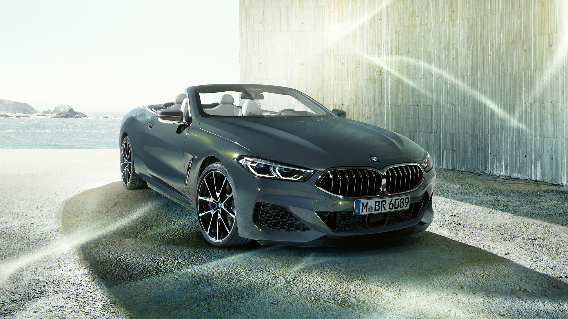 BMW M850i xDrive Кабриолет, вид спереди с поворотом в три четверти. BMW M850i xDrive Кабриолет в Dravit Grey металлик с открытым мягким верхом, вид спереди с поворотом в три четверти, на берегу.