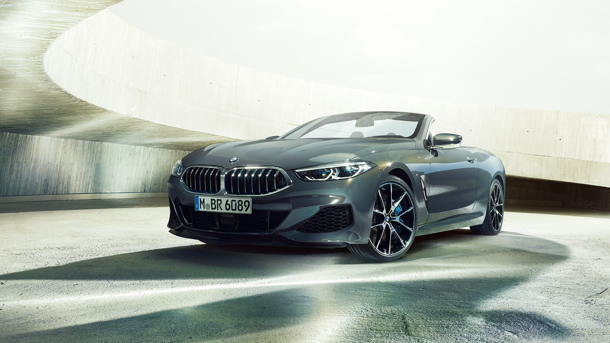 BMW M850i xDrive Кабриолет, вид спереди с поворотом в три четверти. BMW M850i xDrive Кабриолет в Dravit Grey металлик с открытым мягким верхом, вид спереди с поворотом в три четверти, перед бетонным зданием.
