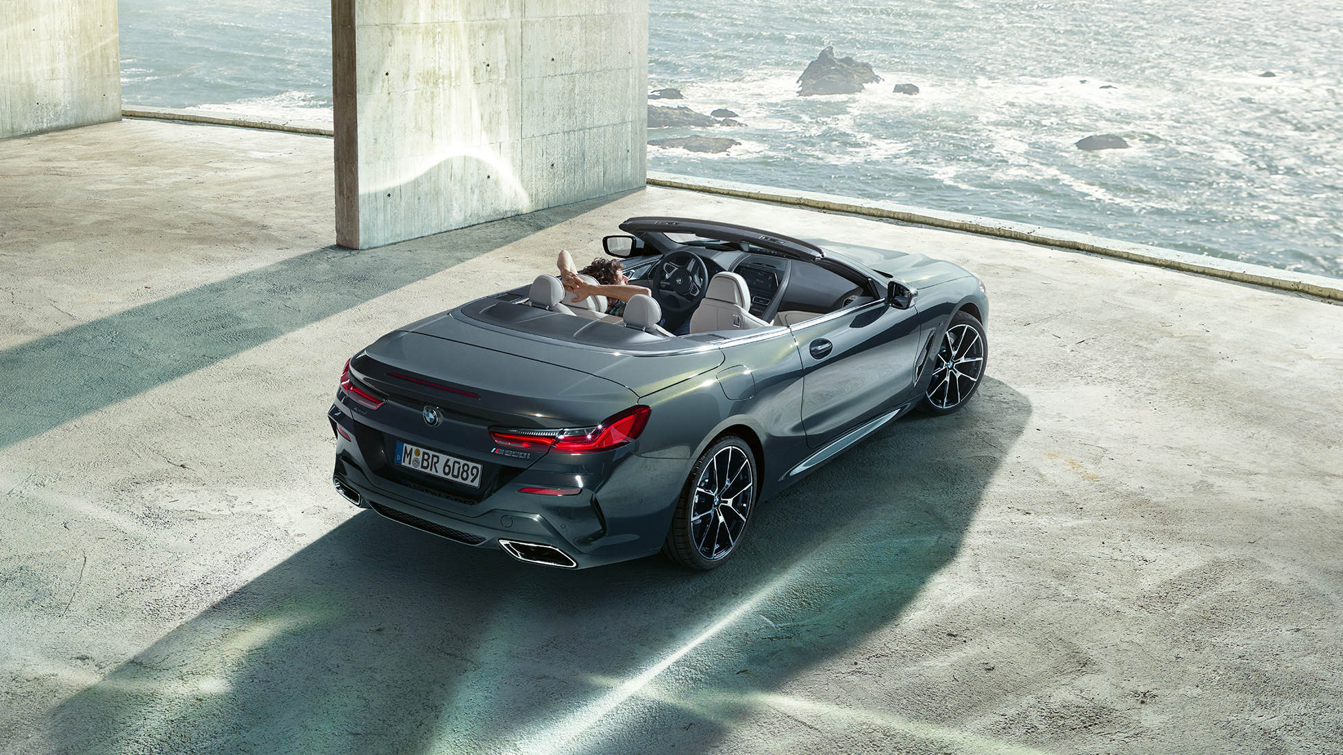 BMW M850i xDrive Кабриолет, с высоты птичьего полета. BMW M850i xDrive Кабриолет в Dravit Grey металлик с открытым мягким верхом, с высоты птичьего полета, парковка в бетонном здании перед океаном.