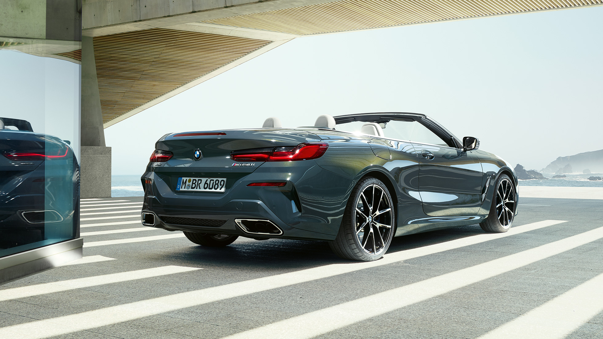 BMW M850i xDrive Кабриолет, вид сзади с поворотом в три четверти. BMW M850i xDrive Кабриолет в Dravit Grey металлик с открытым мягким верхом, вид сзади с поворотом в три четверти, парковка перед океаном.