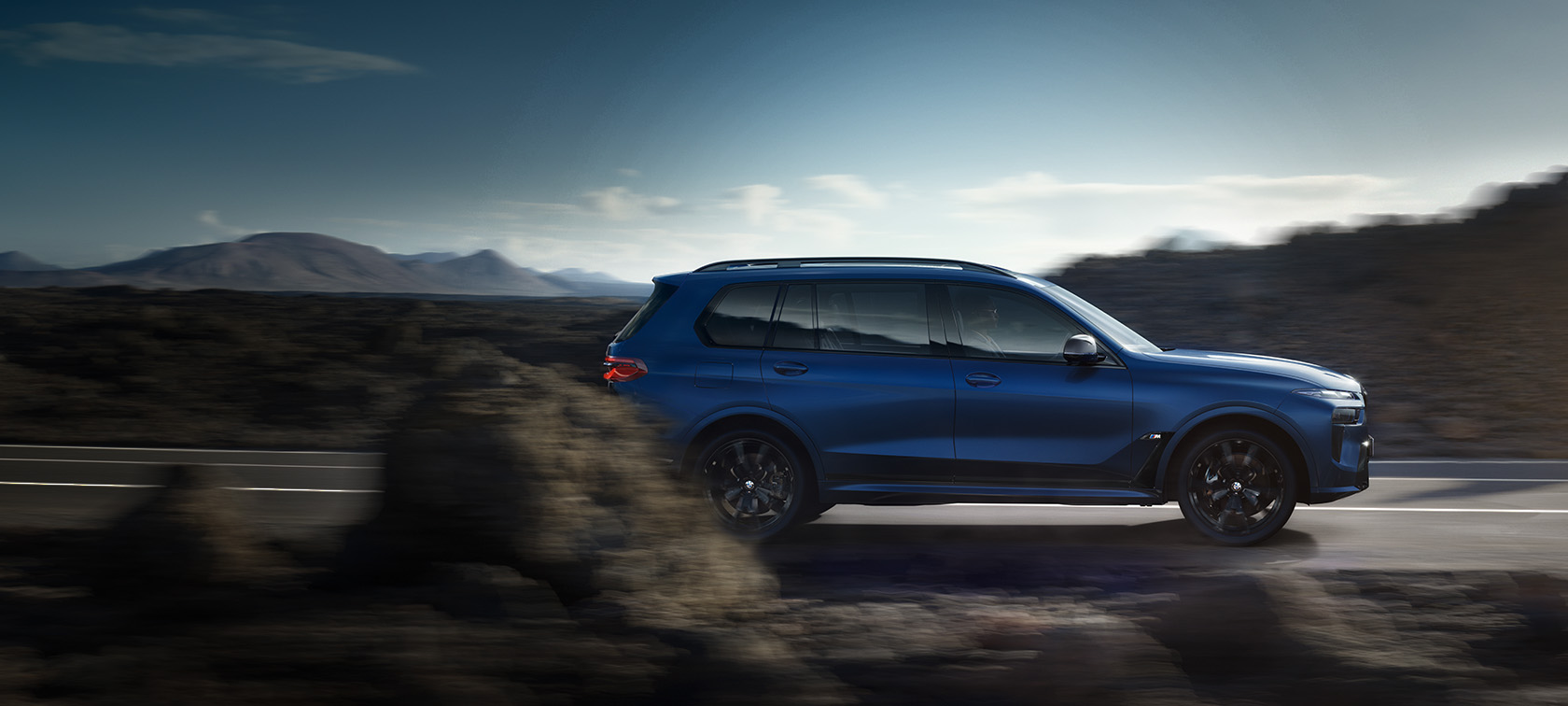 BWM X7 M60i G07 LCI синего цвета Frozen Marina Bay Blue, в движении на улице, вид сбоку BWM X7 M60i G07 LCI синего цвета Frozen Marina Bay Blue, на улице на фоне горного пейзажа, вид сбоку